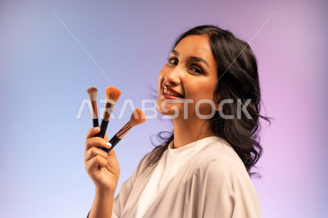 استعمال فرشاة أحمر الخدود, مفهوم التزين والتجمل, بورتريه مقرب من الجانب لامرأة عربية خليجية سعودية ترتدي العباءة تمسك فرش المكياج تنظر إلى الكاميرا بإيماءات النعومة والثقة، الاهتمام والعناية بالمظهر الخارجي, التسويق لأدوات ومنتجات التجميل, خلفية ملونة