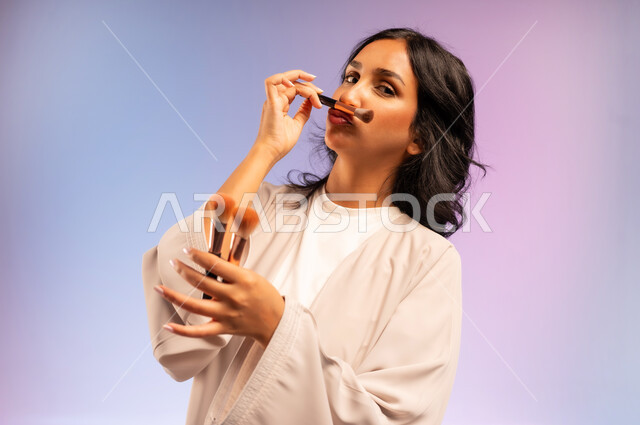 مفهوم التزين والتجمل, بورتريه مقرب لامرأة عربية خليجية سعودية ترتدي العباءة تمسك فرش المكياج تنظر إلى الكاميرا بإيماءات النعومة والثقة، الاهتمام والعناية بالمظهر الخارجي, استعمال فرشاة أحمر الخدود, التسويق لأدوات ومنتجات التجميل, خلفية ملونة