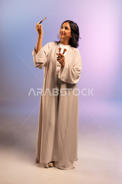 استعمال فرشاة أحمر الخدود, الاهتمام والعناية بالمظهر الخارجي, مفهوم التزين والتجمل, بورتريه لامرأة عربية خليجية سعودية ترتدي العباءة تمسك فرش المكياج تنظر إليها بإيماءات النعومة والسرور، التسويق لأدوات ومنتجات التجميل, الجسم بالطول الكامل، خلفية ملونة