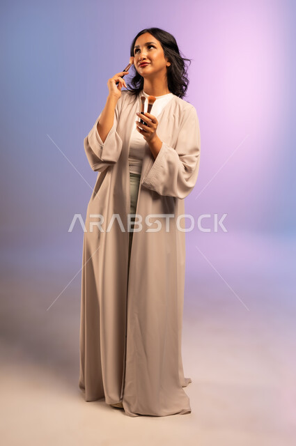 الاهتمام والعناية بالمظهر الخارجي, مفهوم التزين والتجمل, بورتريه لامرأة عربية خليجية سعودية ترتدي العباءة تمسك فرش المكياج تنظر إلى الأعلى بإيماءات النعومة والثقة، استعمال فرشاة أحمر الخدود, التسويق لأدوات ومنتجات التجميل, الجسم بالطول الكامل، خلفية ملونة