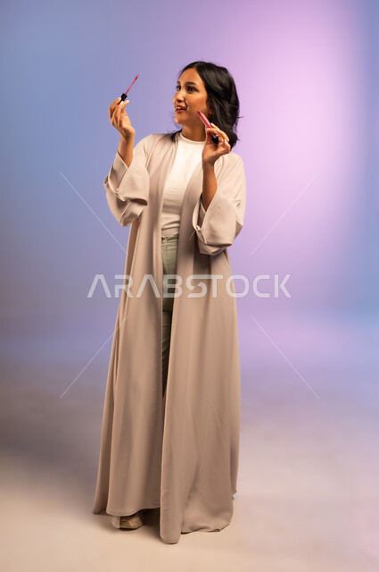 الاهتمام بالجمال والمظهر الخارجي, أداة مكياج تتوريال, بورتريه لامرأة عربية سعودية خليجية ترتدي العباءة تمسك بيدها أحمر الشفاه تنظر إليه، التسويق لمستحضر ومنتج تجميلي نسائي, مفهوم التزين والتجمل, الجسم بالطول الكامل، خلفية ملونة