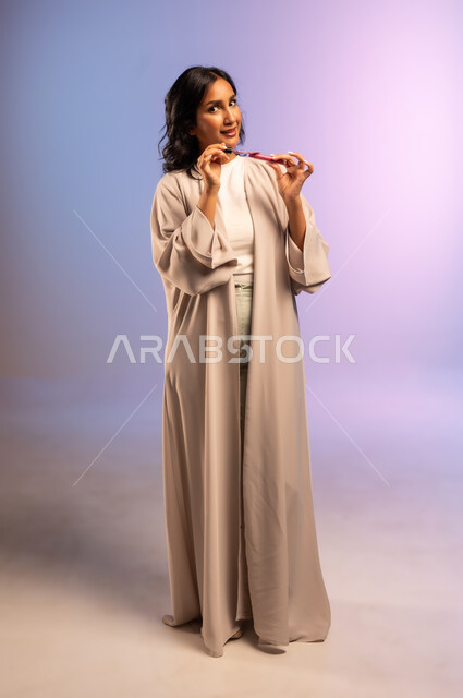 الاهتمام بالجمال والمظهر الخارجي, التسويق لمستحضر ومنتج تجميلي نسائي, أداة مكياج تتوريال, بورتريه لامرأة عربية سعودية خليجية ترتدي العباءة تمسك بيدها أحمر الشفاه تنظر الى الكاميرا بإيماءات السرور والنعومة, مفهوم التزين والتجمل, الجسم بالطول الكامل، خلفية ملونة