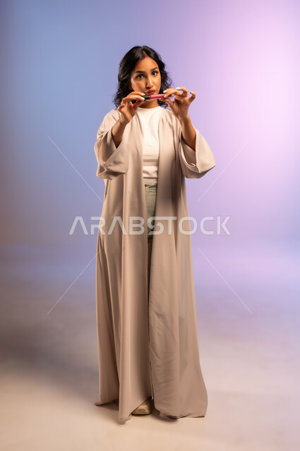 أداة مكياج تتوريال, بورتريه لامرأة عربية سعودية خليجية ترتدي العباءة تمسك بيدها أحمر الشفاه تنظر الى الكاميرا بإيماءات السرور والنعومة, الاهتمام بالجمال والمظهر الخارجي, التسويق لمستحضر ومنتج تجميلي نسائي, مفهوم التزين والتجمل, الجسم بالطول الكامل، خلفية ملونة