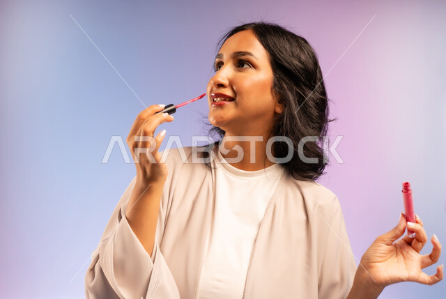 مفهوم التزين والتجمل, التسويق لمستحضر ومنتج تجميلي نسائي, الاهتمام بالجمال والمظهر الخارجي, أداة مكياج تتوريال, بورتريه مقرب لامرأة عربية سعودية خليجية مبتسمة ترتدي العباءة تمسك بيدها أحمر الشفاه تنظر إلى شيء ما في الاعلى بإيماءات السرور، خلفية ملونة