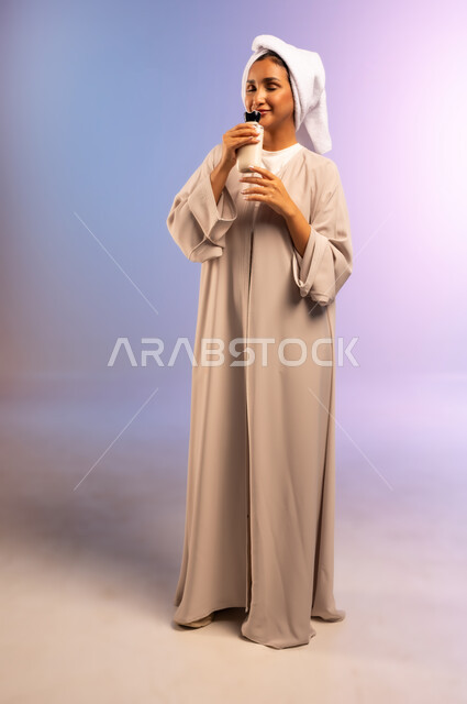 الاهتمام بالأناقة والمظهر الخارجي، مفهوم العناية بنضارة وتحسين البشرة، بورتريه لامرأة عربية خليجية سعودية ترتدي العباءة تضع منشفة بيضاء على رأسها تمسك علبة كريم الترطيب تقوم بشم رائحتها، استخدام مستحضر ومكون تجميلي، الجسم بالطول الكامل، خلفية ملونة