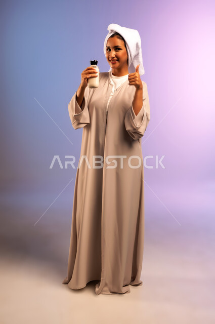 علامة الجودة والامتياز، استخدام مستحضر ومكون تجميلي، مفهوم العناية بنضارة وتحسين البشرة، بورتريه لامرأة عربية خليجية سعودية ترتدي العباءة تضع منشفة بيضاء على رأسها تمسك علبة كريم الترطيب ترفع إصبع الابهام بإيماءات الاعجاب والقبول، الجسم بالطول الكامل، خلفية ملونة