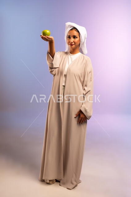 تلبية احيتاجات الجسم من العناصر الغذائية الضرورية, بورتريه لامرأة عربية خليجية سعودية ترتدي العباءة تضع منشفة بيضاء على رأسها تمسك تفاحة خضراء، اتباع نظام غذائي صحي، فطور وسناك لذيذ، أهمية وفائدة الفواكه الطازجة للبشرة، الجسم بالطول الكامل، خلفية ملونة