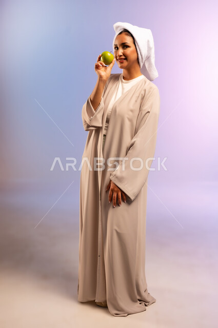 فطور وسناك لذيذ، تلبية احيتاجات الجسم من العناصر الغذائية الضرورية, بورتريه لامرأة عربية خليجية سعودية ترتدي العباءة تضع منشفة بيضاء على رأسها تمسك تفاحة خضراء، اتباع نظام غذائي صحي، أهمية وفائدة الفواكه الطازجة للبشرة، الجسم بالطول الكامل، خلفية ملونة