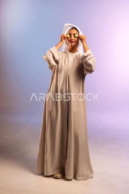 أهمية الخضار الطازجة للبشرة، مفهوم الراحة والاسترخاء، تلبية حاجة الجسم من العناصر الغذائية, بورتريه لامرأة عربية خليجية سعودية ترتدي العباءة تضع منشفة بيضاء على رأسها وشرائح من الخيار على عينيها، اتباع نظام غذائي صحي، الجسم بالطول الكامل، خلفية ملونة