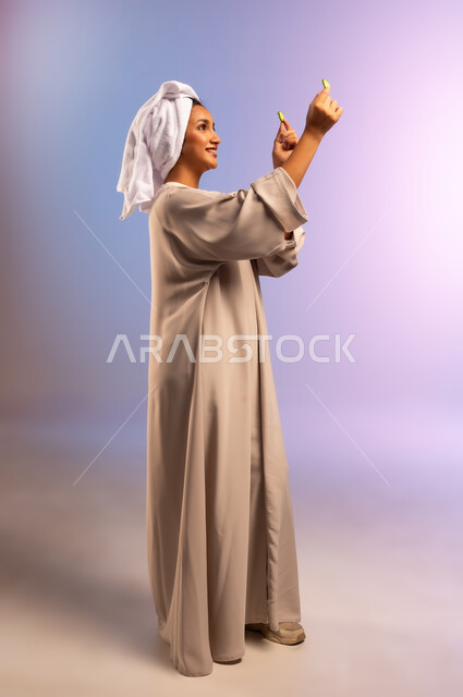مفهوم الراحة والاسترخاء، تلبية حاجة الجسم من العناصر الغذائية, بورتريه من الجانب لامرأة عربية خليجية سعودية ترتدي العباءة تضع منشفة بيضاء على رأسها تمسك بيديها شرائح من الخيار، اتباع نظام غذائي صحي، أهمية الخضار الطازجة للبشرة، الجسم بالطول الكامل، خلفية ملونة