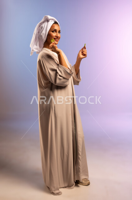 أهمية الخضار الطازجة للبشرة، مفهوم الراحة والاسترخاء، تلبية حاجة الجسم من العناصر الغذائية, بورتريه من الجانب لامرأة عربية خليجية سعودية ترتدي العباءة تضع منشفة بيضاء على رأسها تمسك بيديها شرائح من الخيار، اتباع نظام غذائي صحي، الجسم بالطول الكامل، خلفية ملونة