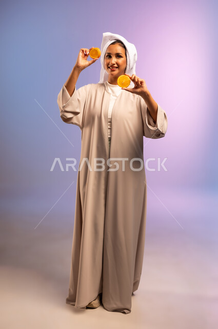 تلبية حاجة الجسم من العناصر الغذائية الضرورية, بورتريه لامرأة عربية خليجية سعودية ترتدي العباءة تضع منشفة بيضاء على رأسها تمسك بيديها شرائح من البرتقال، أهمية الفواكه الطازجة للبشرة، اتباع نظام غذائي صحي، فطور وسناك لذيذ، الجسم بالطول الكامل، خلفية ملونة