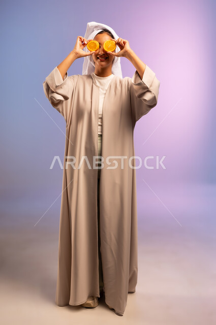 فطور وسناك لذيذ، اتباع نظام غذائي صحي، تلبية حاجة الجسم من العناصر الغذائية, بورتريه لامرأة عربية خليجية سعودية ترتدي العباءة تضع منشفة بيضاء على رأسها تمسك بيديها شرائح من البرتقال تضعها أمام عينيها، أهمية الفواكه الطازجة للبشرة، الجسم بالطول الكامل، خلفية ملونة