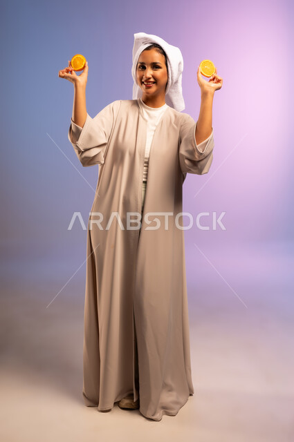 تلبية حاجة الجسم من العناصر الغذائية الضرورية, بورتريه لامرأة عربية خليجية سعودية ترتدي العباءة تضع منشفة بيضاء على رأسها تمسك بيديها شرائح من البرتقال، أهمية الفواكه الطازجة للبشرة، اتباع نظام غذائي صحي، فطور وسناك لذيذ، الجسم بالطول الكامل، خلفية ملونة