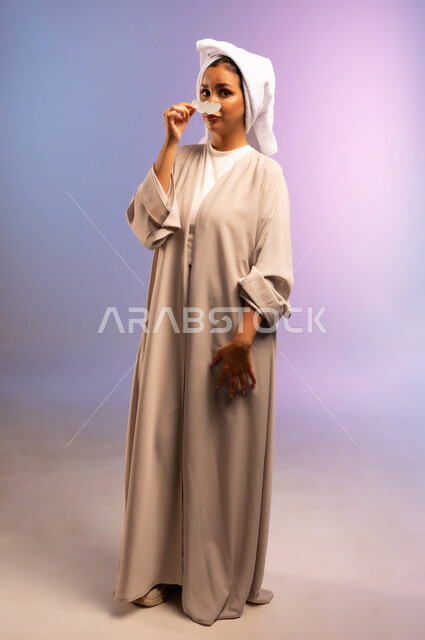 استخدام ماسك تنظيف المسام، التخلص من الرؤوس السوداء والجلد الميت، بورتريه لامرأة عربية خليجية سعودية ترتدي العباءة تضع منشفة بيضاء على رأسها تقوم بوضع لصقات الأنف، العناية بنضارة وتحسين البشرة، مفهوم الاهتمام بالمظهر الخارجي، الجسم بالطول الكامل، خلفية ملونة