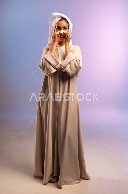 العناية بنضارة وتحسين البشرة، استخدام ماسك تنظيف المسام، التخلص من الرؤوس السوداء والجلد الميت، بورتريه لامرأة عربية خليجية سعودية ترتدي العباءة تضع منشفة بيضاء على رأسها تقوم بوضع لصقات الأنف، مفهوم الاهتمام بالمظهر الخارجي، الجسم بالطول الكامل، خلفية ملونة