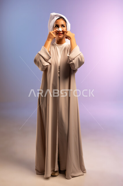 استخدام ماسك تنظيف المسام، التخلص من الرؤوس السوداء والجلد الميت، بورتريه لامرأة عربية خليجية سعودية ترتدي العباءة تضع منشفة بيضاء على رأسها تقوم بوضع لصقات الأنف، العناية بنضارة وتحسين البشرة، مفهوم الاهتمام بالمظهر الخارجي، الجسم بالطول الكامل، خلفية ملونة