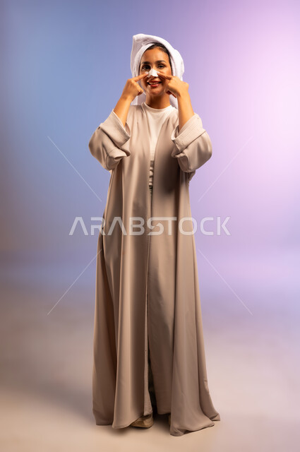 العناية بنضارة وتحسين البشرة، استخدام ماسك تنظيف المسام، التخلص من الرؤوس السوداء والجلد الميت، بورتريه لامرأة عربية خليجية سعودية ترتدي العباءة تضع منشفة بيضاء على رأسها تقوم بوضع لصقات الأنف، مفهوم الاهتمام بالمظهر الخارجي، الجسم بالطول الكامل، خلفية ملونة