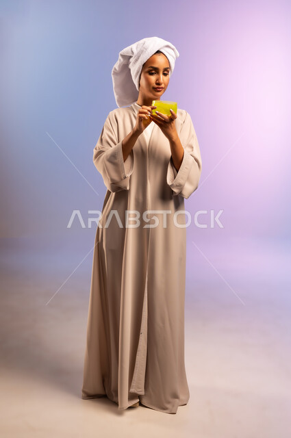 الاهتمام بالأناقة والمظهر الخارجي، مفهوم العناية بنضارة وتحسين البشرة، بورتريه لامرأة عربية خليجية سعودية ترتدي العباءة تضع منشفة بيضاء على رأسها تمسك علبة كريم الترطيب، استخدام مستحضر ومكون تجميلي، الجسم بالطول الكامل، خلفية ملونة