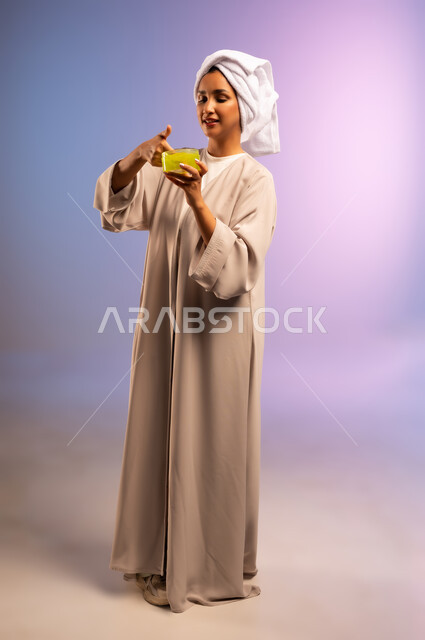 الاهتمام بالأناقة والمظهر الخارجي، مفهوم العناية بنضارة وتحسين البشرة، بورتريه لامرأة عربية خليجية سعودية ترتدي العباءة تضع منشفة بيضاء على رأسها تمسك علبة كريم الترطيب، استخدام مستحضر ومكون تجميلي، الجسم بالطول الكامل، خلفية ملونة