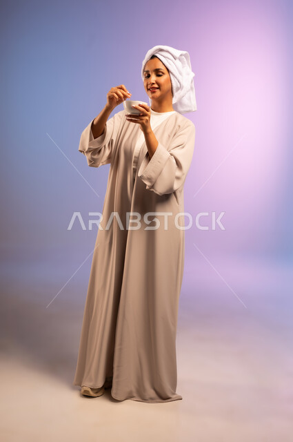 مفهوم الاهتمام بالمظهر الخارجي عند النساء، استخدام ماسك بمكونات طبيعية للتنظيف والتقشير، بورتريه لامرأة عربية خليجية سعودية ترتدي العباءة تضع منشفة بيضاء على رأسها تمسك وعاء فيه خلطة تجميلية منزلية، المحافظة على نضارة البشرة، الجسم بالطول الكامل، خلفية ملونة