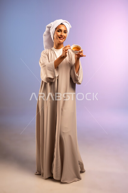 مفهوم الاهتمام بالمظهر الخارجي عند النساء، استخدام ماسك بمكونات طبيعية للتنظيف والتقشير، بورتريه لامرأة عربية خليجية سعودية ترتدي العباءة تضع منشفة بيضاء على رأسها تمسك وعاء فيه خلطة تجميلية منزلية، المحافظة على نضارة البشرة، الجسم بالطول الكامل، خلفية ملونة