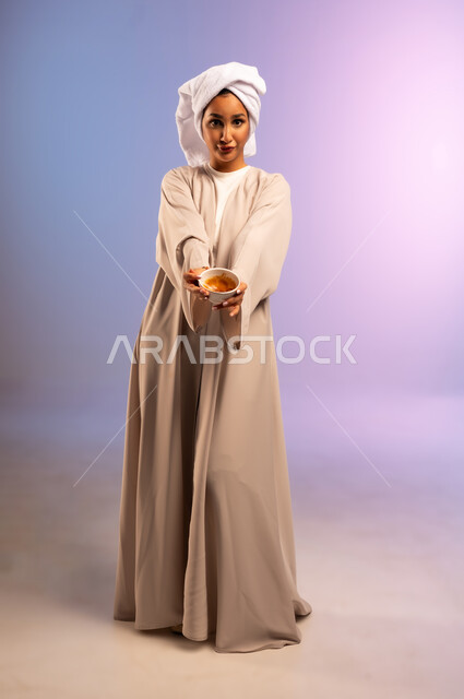 استخدام ماسك بمكونات طبيعية للتنظيف والتقشير، بورتريه لامرأة عربية خليجية سعودية ترتدي العباءة تضع منشفة بيضاء على رأسها تمسك وعاء فيه خلطة تجميلية منزلية، المحافظة على نضارة البشرة، مفهوم الاهتمام بالمظهر الخارجي عند النساء، الجسم بالطول الكامل، خلفية ملونة
