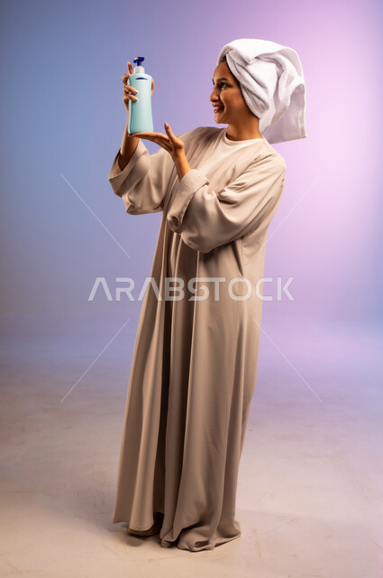 الاهتمام بالأناقة والمظهر الخارجي، مفهوم العناية بنضارة وتحسين البشرة، بورتريه لامرأة عربية خليجية سعودية ترتدي العباءة تضع منشفة بيضاء على رأسها تمسك علبة كريم الترطيب، استخدام مستحضر ومكون تجميلي، الجسم بالطول الكامل، خلفية ملونة