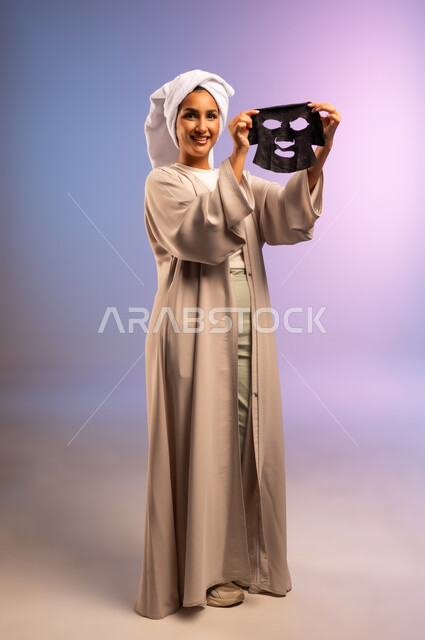 استخدام مكون ومستحضر تجميلي للتنظيف والتقشير، بورتريه لامرأة عربية خليجية سعودية ترتدي العباءة تضع منشفة بيضاء على رأسها تمسك قناع الطين تستعد لوضعه على وجهها، المحافظة على نضارة البشرة، مفهوم الاهتمام بالمظهر الخارجي عند النساء، الجسم بالطول الكامل، خلفية ملونة