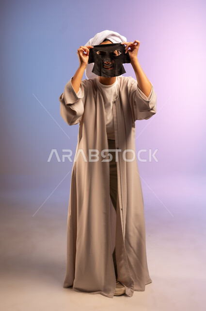 المحافظة على نضارة البشرة، استخدام مكون ومستحضر تجميلي للتنظيف والتقشير، بورتريه لامرأة عربية خليجية سعودية ترتدي العباءة تضع منشفة بيضاء على رأسها تقوم بوضع قناع الطين على وجهها، مفهوم الاهتمام بالمظهر الخارجي عند النساء، الجسم بالطول الكامل، خلفية ملونة