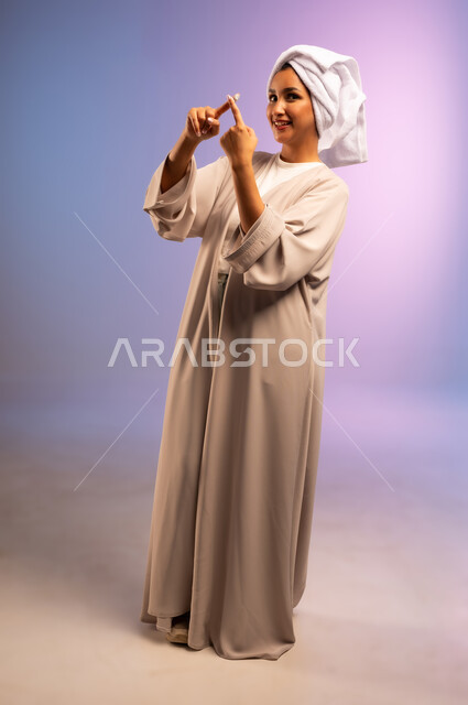 الاهتمام بالمظهر الخارجي، استخدام مستحضر ومكون تجميلي، مفهوم العناية بنضارة وتحسين البشرة، بورتريه لامرأة عربية خليجية سعودية ترتدي العباءة تضع منشفة بيضاء على رأسها تضع كريم الترطيب على إصبعيها تنظر إلى الكاميرا بإيماءات السرور، الجسم بالطول الكامل، خلفية ملونة