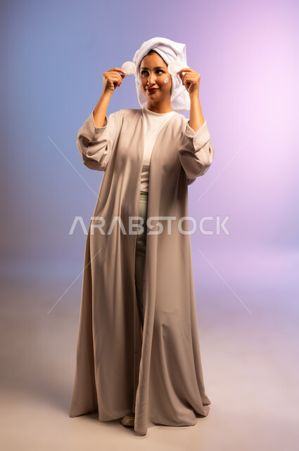 إزالة المكياج باستخدام ضمادات قطنية، الاهتمام بالمظهر الخارجي، مفهوم العناية بنضارة ونظافة البشرة، بورتريه لامرأة عربية خليجية سعودية ترتدي العباءة تضع منشفة بيضاء على رأسها تمسك بيديها قطنتين تقوم بمسح وجهها، الجسم بالطول الكامل، خلفية ملونة