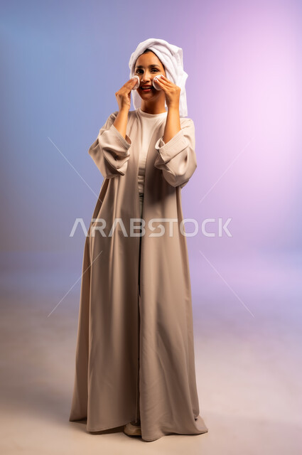 الاهتمام بالمظهر الخارجي، مفهوم العناية بنضارة ونظافة البشرة، بورتريه لامرأة عربية خليجية سعودية ترتدي العباءة تضع منشفة بيضاء على رأسها تمسك بيديها قطنتين تقوم بمسح وجهها، إزالة المكياج باستخدام ضمادات قطنية، الجسم بالطول الكامل، خلفية ملونة