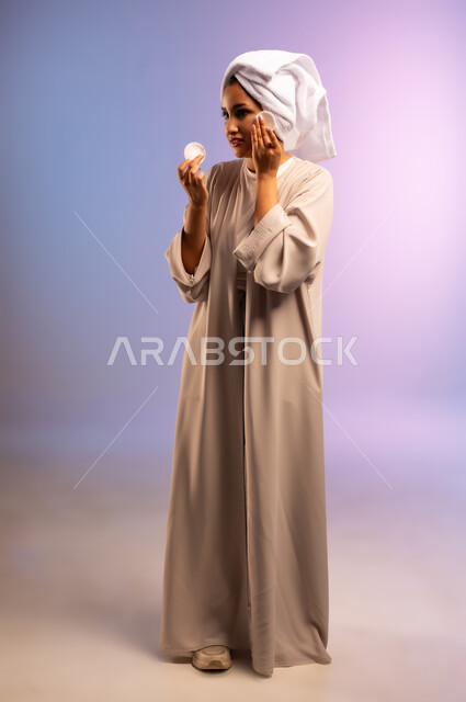 إزالة المكياج باستخدام ضمادات قطنية، الاهتمام بالمظهر الخارجي، مفهوم العناية بنضارة ونظافة البشرة، بورتريه لامرأة عربية خليجية سعودية ترتدي العباءة تضع منشفة بيضاء على رأسها تمسك بيديها قطنتين تقوم بمسح وجهها، الجسم بالطول الكامل، خلفية ملونة