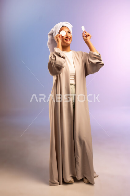 إزالة المكياج باستخدام ضمادات قطنية، الاهتمام بالمظهر الخارجي، مفهوم العناية بنضارة ونظافة البشرة، بورتريه لامرأة عربية خليجية سعودية ترتدي العباءة تضع منشفة بيضاء على رأسها تمسك بيديها قطنتين تقوم بمسح وجهها، الجسم بالطول الكامل، خلفية ملونة