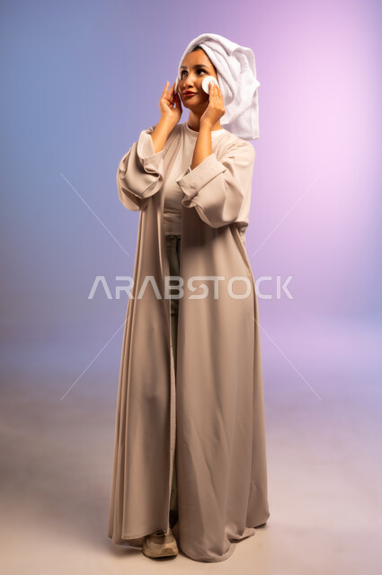 الاهتمام بالمظهر الخارجي، مفهوم العناية بنضارة ونظافة البشرة، بورتريه لامرأة عربية خليجية سعودية ترتدي العباءة تضع منشفة بيضاء على رأسها تمسك بيديها قطنتين تقوم بمسح وجهها، إزالة المكياج باستخدام ضمادات قطنية، الجسم بالطول الكامل، خلفية ملونة