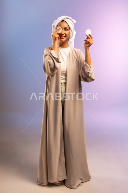 الاهتمام بالمظهر الخارجي، مفهوم العناية بنضارة ونظافة البشرة، بورتريه لامرأة عربية خليجية سعودية ترتدي العباءة تضع منشفة بيضاء على رأسها تمسك بيديها قطنتين تقوم بمسح وجهها، إزالة المكياج باستخدام ضمادات قطنية، الجسم بالطول الكامل، خلفية ملونة