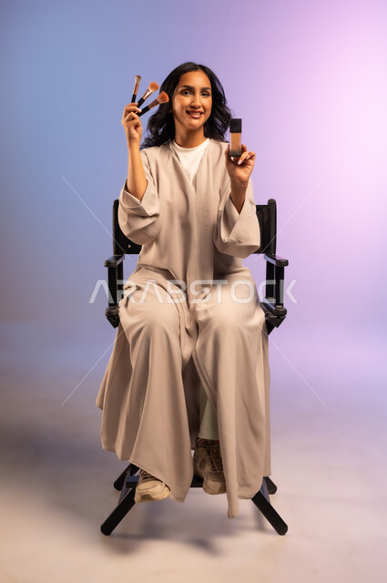 استخدام المسحوق التجميلي, مفهوم التزين والتجمل, بورتريه لامرأة عربية خليجية سعودية ترتدي العباءة تجلس على الكرسي تمسك فرش المكياج ومستحضر للوجه، الاهتمام والعناية بالمظهر الخارجي, استعمال فرشاة أحمر الخدود, التسويق لمستحضرات ومنتجات التجميل, خلفية ملونة