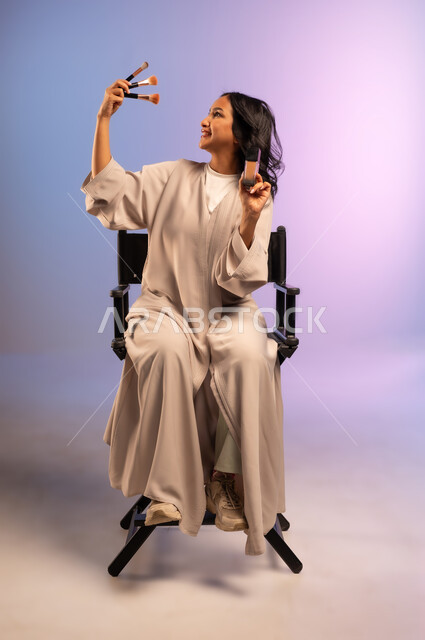 استعمال فرشاة أحمر الخدود, الاهتمام والعناية بالمظهر الخارجي, استخدام المسحوق التجميلي, مفهوم التزين والتجمل, بورتريه لامرأة عربية خليجية سعودية ترتدي العباءة تجلس على الكرسي تمسك فرش المكياج ومستحضر للوجه، التسويق لمستحضرات ومنتجات التجميل, خلفية ملونة