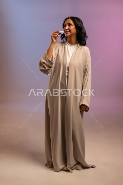 الحصول على ابتسامة هوليوود باستخدام التقنية الحديثة، العناية والاهتمام بصحة ونظافة الفم، بورتريه لامرأة عربية خليجية سعودية ترتدي العباءة تضع منشفة بيضاء على رأسها تستخدم جهاز تبييض الأسنان الليزري المنزلي، الجسم بالطول الكامل، خلفية ملونة