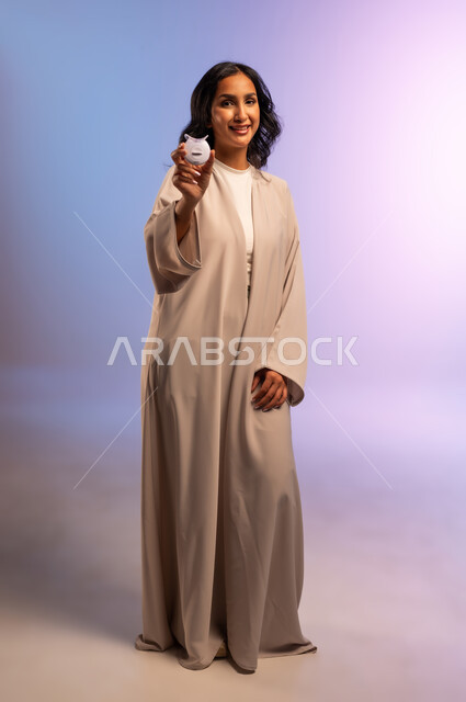 Get a Hollywood smile using modern technology, care and attention to oral health and hygiene, portrait of a Saudi Arabian Gulf woman wearing an abaya, putting a white towel on her head, holding a home laser teeth whitening device in her hand, looking at the camera, full body, colored background