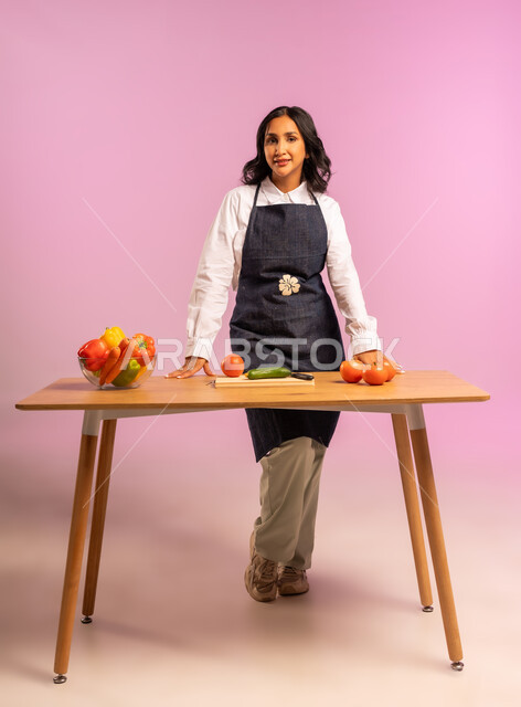 اتباع نظام غذائي نباتي، تجهيز مكونات السلطة، مفهوم الأكل الصحي، بورتريه لامرأة عربية خليجية سعودية ترتدي المئزر تقف خلف طاولة خشبية تنظر إلى الكاميرا بإيماءات الثقة بالنفس، تحضير السلطات والمأكولات الشهية، وجبة طعام صحية، الجسم بالطول الكامل، خلفية ملونة
