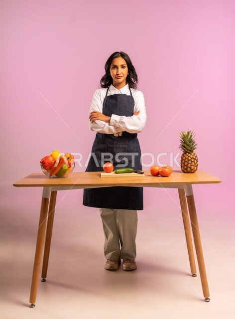 تجهيز مكونات السلطة، مفهوم الأكل الصحي، بورتريه لامرأة عربية خليجية سعودية ترتدي المئزر تقف خلف طاولة خشبية بأيجي متقاطعة تنظر إلى الكاميرا بإيماءات الثقة بالنفس، اتباع نظام غذائي نباتي، تحضير السلطات الشهية، وجبة طعام صحية، الجسم بالطول الكامل، خلفية ملونة