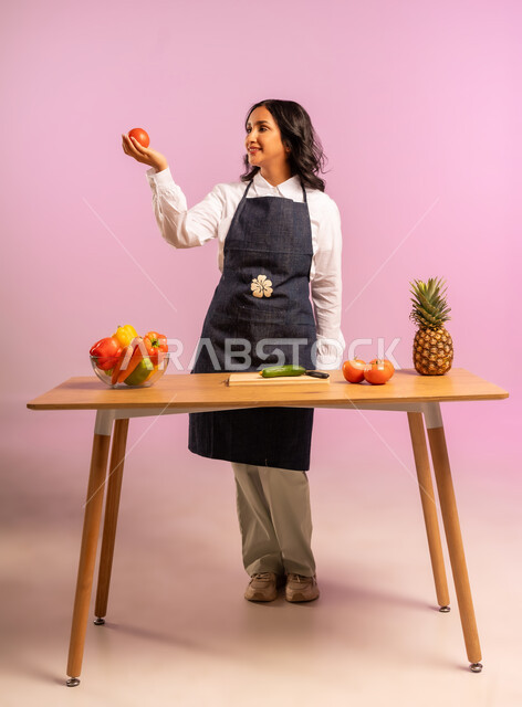 التجهيز لاعداد السلطة، مفهوم الأكل الصحي، بورتريه لامرأة عربية خليجية سعودية ترتدي المئزر تقف خلف طاولة خشبية تحمل حبة طماطم تنظر إليها، الاستمتاع بتحضير السلطات والمأكولات الشهية، وجبة طعام صحية، اتباع نظام غذائي نباتي، الجسم بالطول الكامل، خلفية ملونة