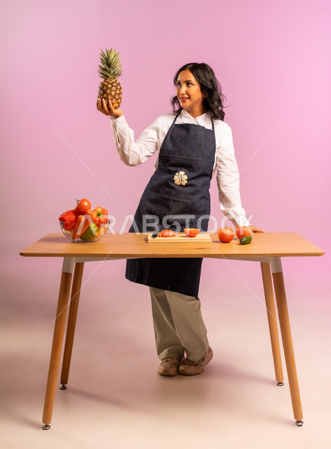 الاستمتاع باعداد السلطة على طاولة المطبخ، اتباع نظام غذائي نباتي، مفهوم الأكل الصحي، بورتريه لامرأة عربية خليجية سعودية ترتدي المئزر تحمل حبة أناناس تنظر إليها، تحضير السلطات والمأكولات الشهية، وجبة طعام صحية، الجسم بالطول الكامل، خلفية ملونة