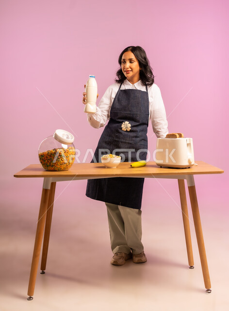 تحضير رقائق الذرة في طبق أبيض، وجبة طعام صحية، مفهوم الأكل الصحي، اتباع نظام غذائي، بورتريه لامرأة عربية خليجية سعودية ترتدي اليونفر تقوم بحمل علبة الحليب و اعداد الفطور الصباحي على طاولة المطبخ، الجسم بالطول الكامل، خلفية ملونة