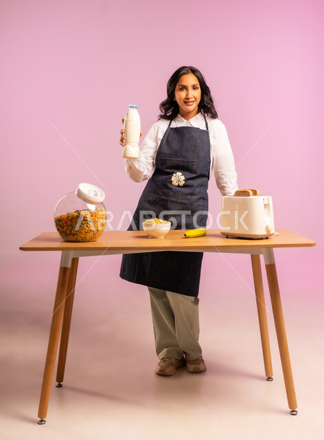 وجبة طعام صحية، تحضير رقائق الذرة في طبق أبيض، مفهوم الأكل الصحي، اتباع نظام غذائي، بورتريه لامرأة عربية خليجية سعودية ترتدي اليونفر تقوم بحمل علبة الحليب و اعداد الفطور الصباحي على طاولة المطبخ، الجسم بالطول الكامل، خلفية ملونة