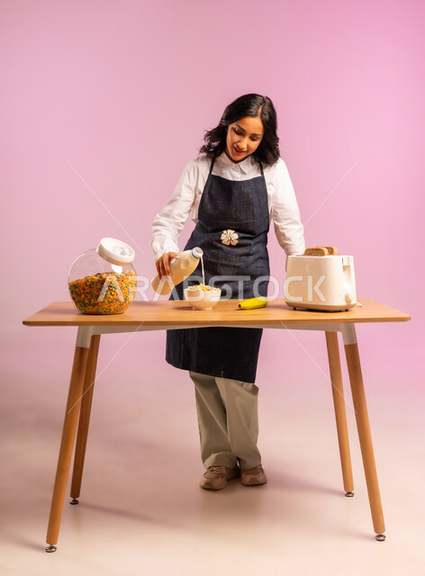 مفهوم الأكل الصحي،  تحضير رقائق الذرة في طبق أبيض بايماءات السعادة والسرور، اتباع نظام غذائي، بورتريه لامرأة عربية خليجية سعودية ترتدي اليونفر تقوم بإعداد الفطور الصباحي على طاولة المطبخ،  وجبة طعام صحية، سكب الحليب فوق حبوب الإفطار، الجسم بالطول الكامل، خلفية ملونة