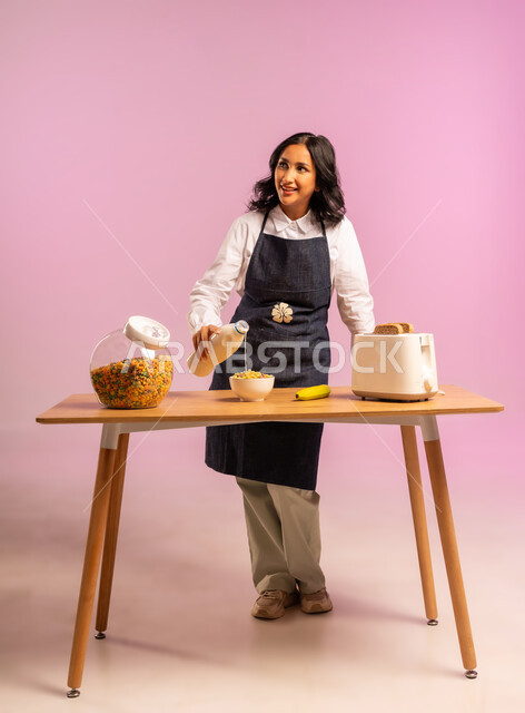 سكب الحليب فوق حبوب الإفطار، مفهوم الأكل الصحي،  تحضير رقائق الذرة في طبق أبيض بايماءات السعادة والسرور، اتباع نظام غذائي، بورتريه لامرأة عربية خليجية سعودية ترتدي اليونفر تقوم بإعداد الفطور الصباحي على طاولة المطبخ،  وجبة طعام صحية، الجسم بالطول الكامل، خلفية ملونة