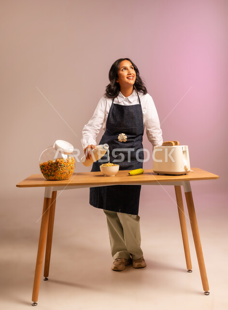 سكب الحليب فوق حبوب الإفطار، مفهوم الأكل الصحي،  تحضير رقائق الذرة في طبق أبيض بايماءات السعادة والسرور، اتباع نظام غذائي، بورتريه لامرأة عربية خليجية سعودية ترتدي اليونفر تقوم بإعداد الفطور الصباحي على طاولة المطبخ،  وجبة طعام صحية، الجسم بالطول الكامل، خلفية ملونة